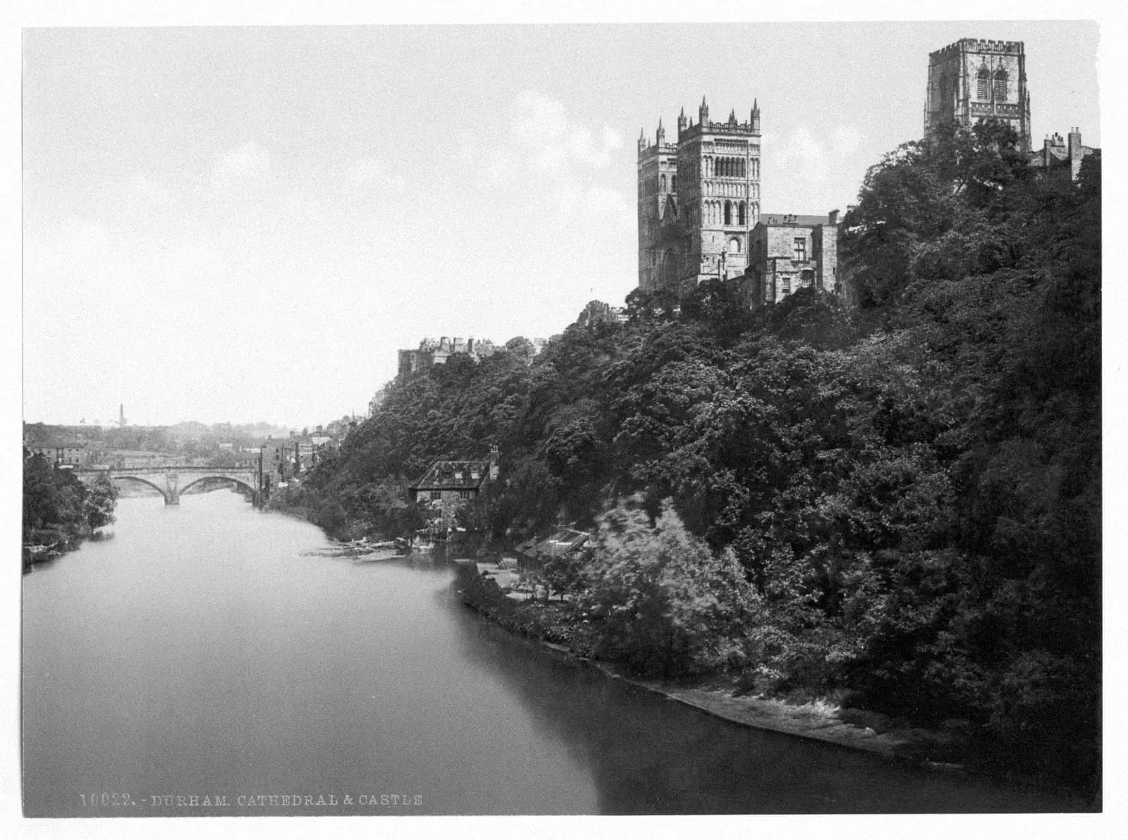 Durham Cathedral's towers rise above the wooded banks of the River Wear, with a stone bridge in the distance.