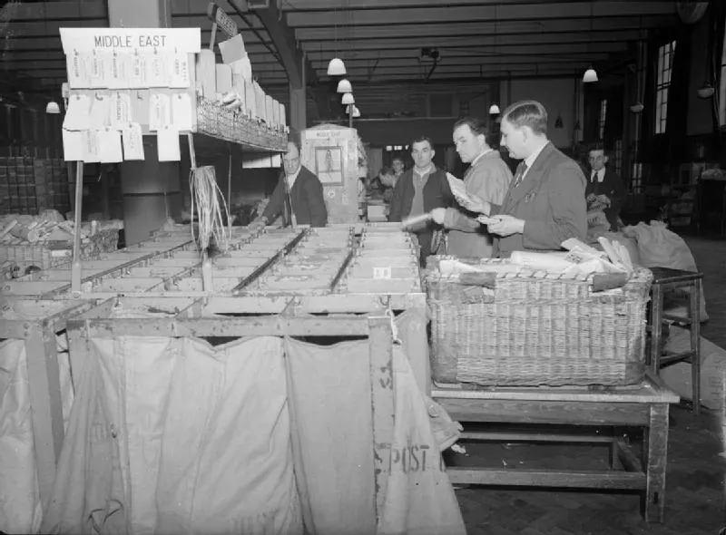 Post Office staff sort packets from wicker baskets into a long frame of hanging mail bags under labelled signs.