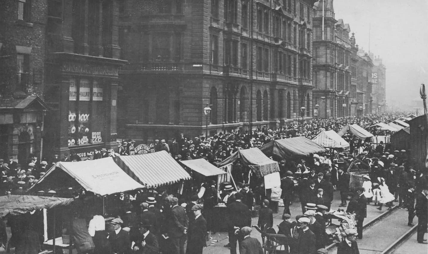 Crowds and canvas market stalls fill Newcastle Quayside on a Sunday morning, below tall stone office buildings.