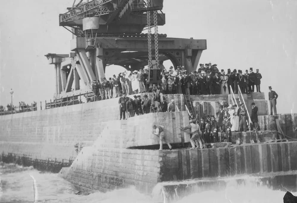 Workmen guide a huge stone block into place on Roker Pier while onlookers in top hats watch from the crane platform above.