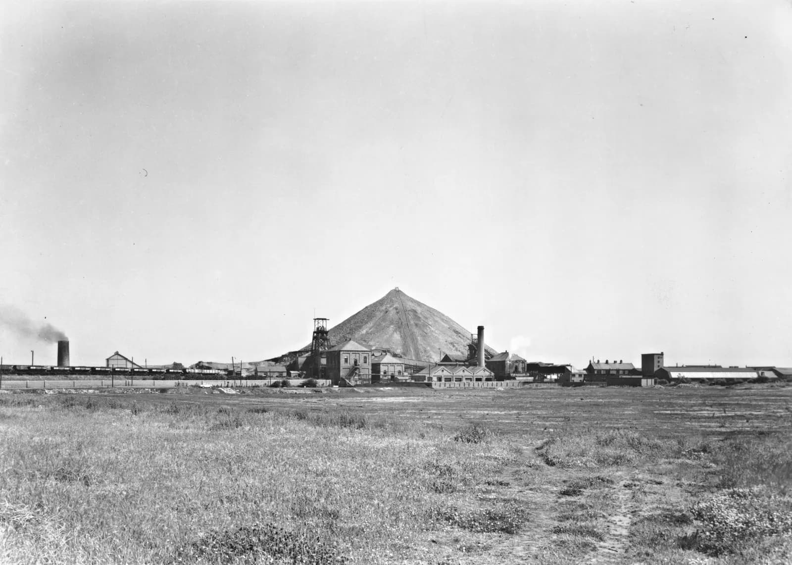 Washington F Pit seen across open fields, with its headgear, chimney and a tall conical spoil heap against a pale sky.